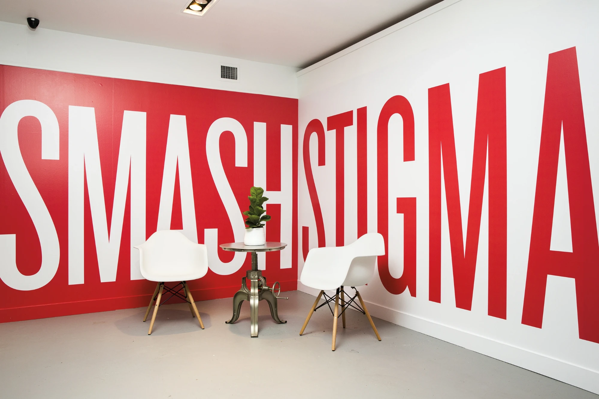 A room with SMASH STIGMA painted floor to ceiling in red and white across two walls, with two white chairs and a small table.