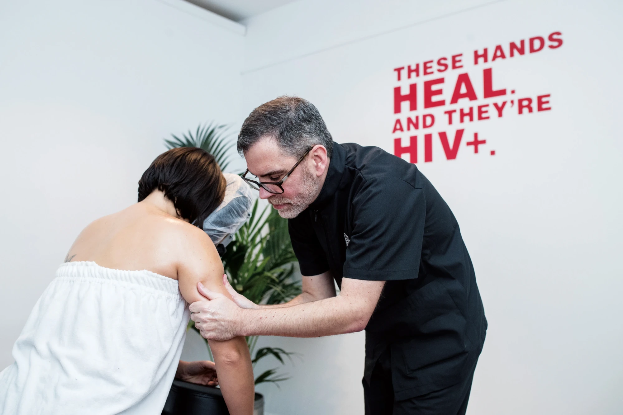 At Healing House, an HIV+ massage therapist works on a client&rsquo;s arm beneath the wall text &ldquo;These hands heal. And they&rsquo;re HIV+.&rdquo;