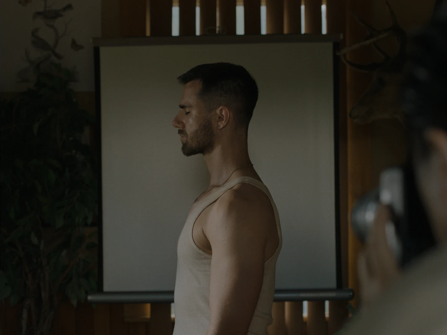 A still from the horror film Others: a man in a tank top stands in profile with his eyes closed as someone photographs him.