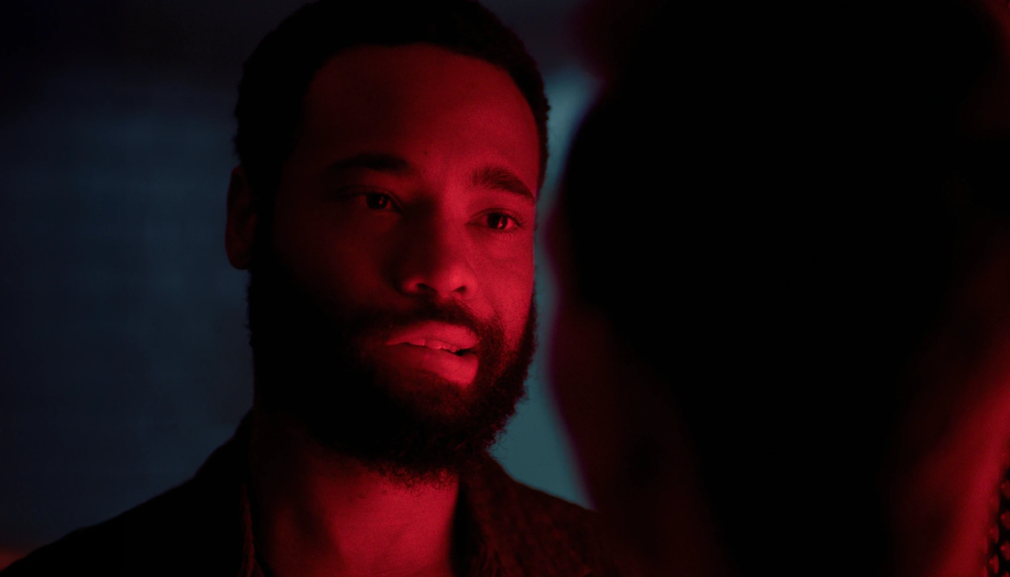 A still from Big Fucking Deal: a man lit in red looks at someone just out of frame.