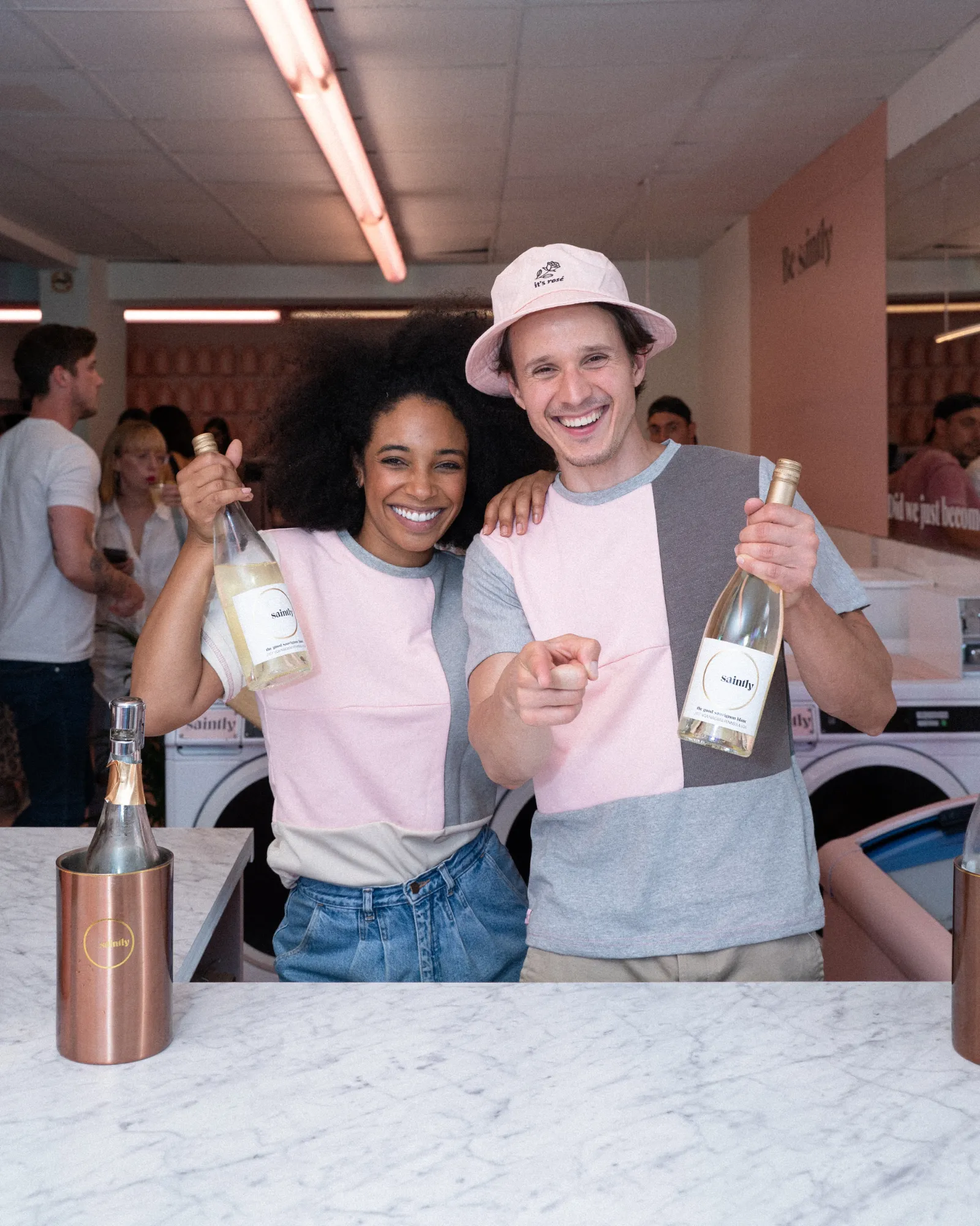 Two smiling hosts in pink-and-gray Saintly T-shirts, one in an &ldquo;it&rsquo;s ros&eacute;&rdquo; bucket hat, hold up bottles of Saintly at a marble bar inside the pink Sip &rsquo;n&rsquo; Suds laundromat.