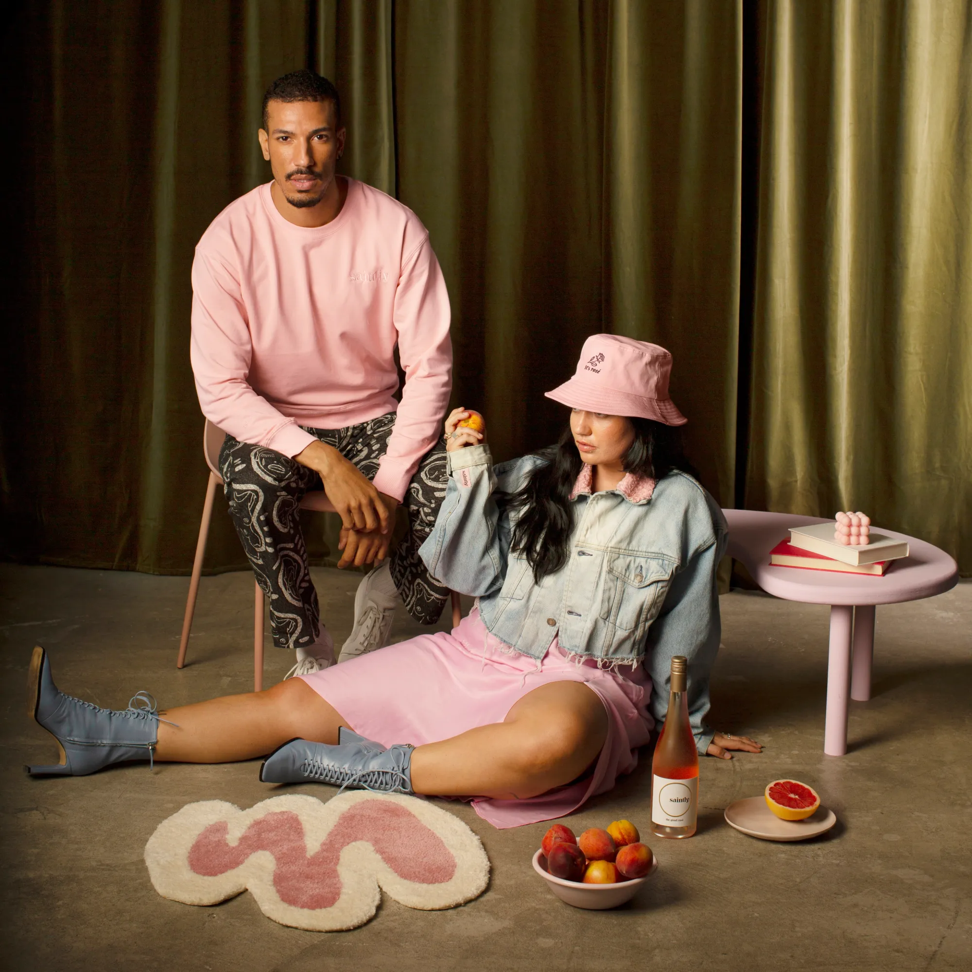 Two people in Saintly merch, a pink sweatshirt and a pink bucket hat, with a bottle of Saintly, peaches and grapefruit, in front of an olive velvet curtain.