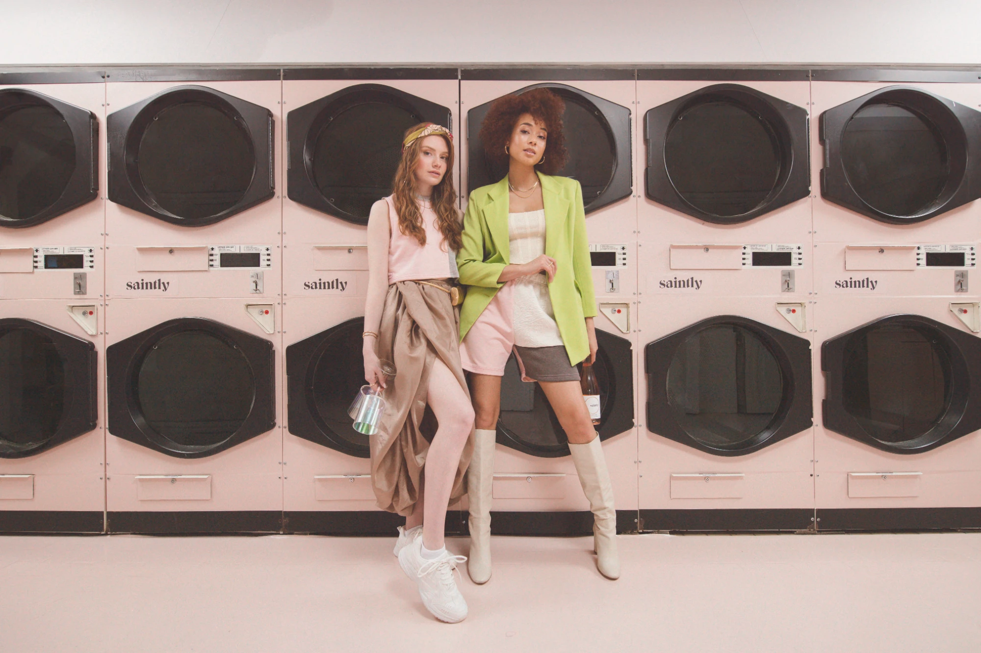 Two women, one in a lime blazer, lean against a wall of Saintly-branded dryers in a pink laundromat.
