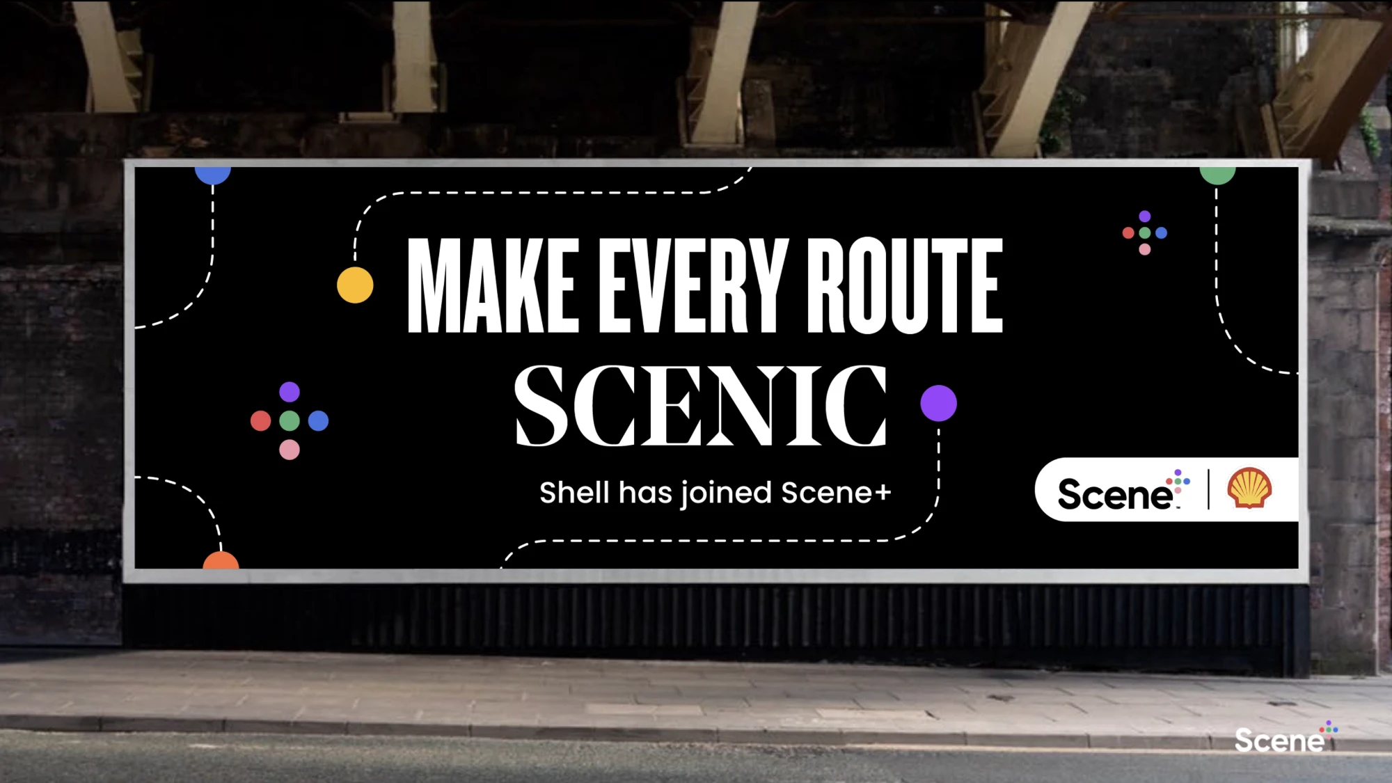 A street billboard under a rail bridge reads &ldquo;Make every route scenic. Shell has joined Scene+&rdquo;, with the Scene+ and Shell logos.