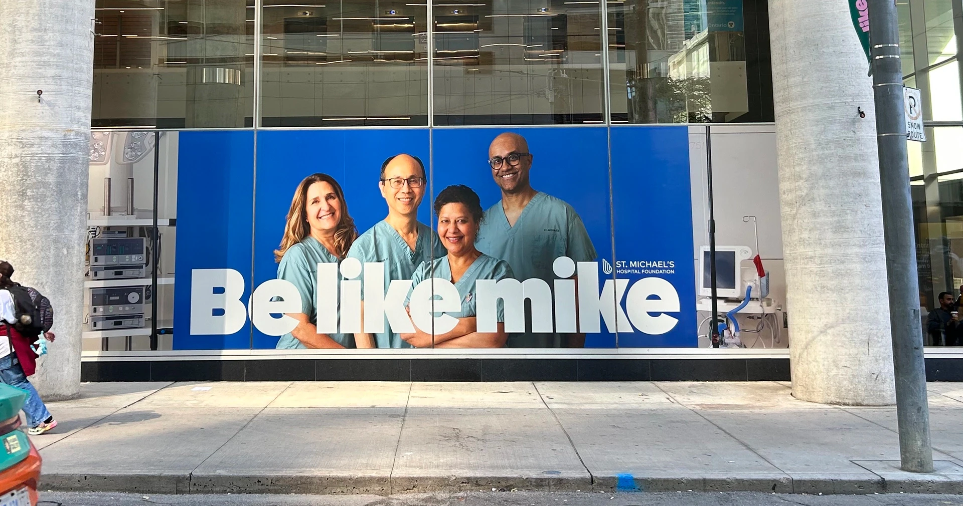 A street-level window at St. Michael&rsquo;s Hospital covered by a &ldquo;Be like mike&rdquo; banner of four smiling staff in scrubs, with the St. Michael&rsquo;s Hospital Foundation logo, as a passer-by walks past.