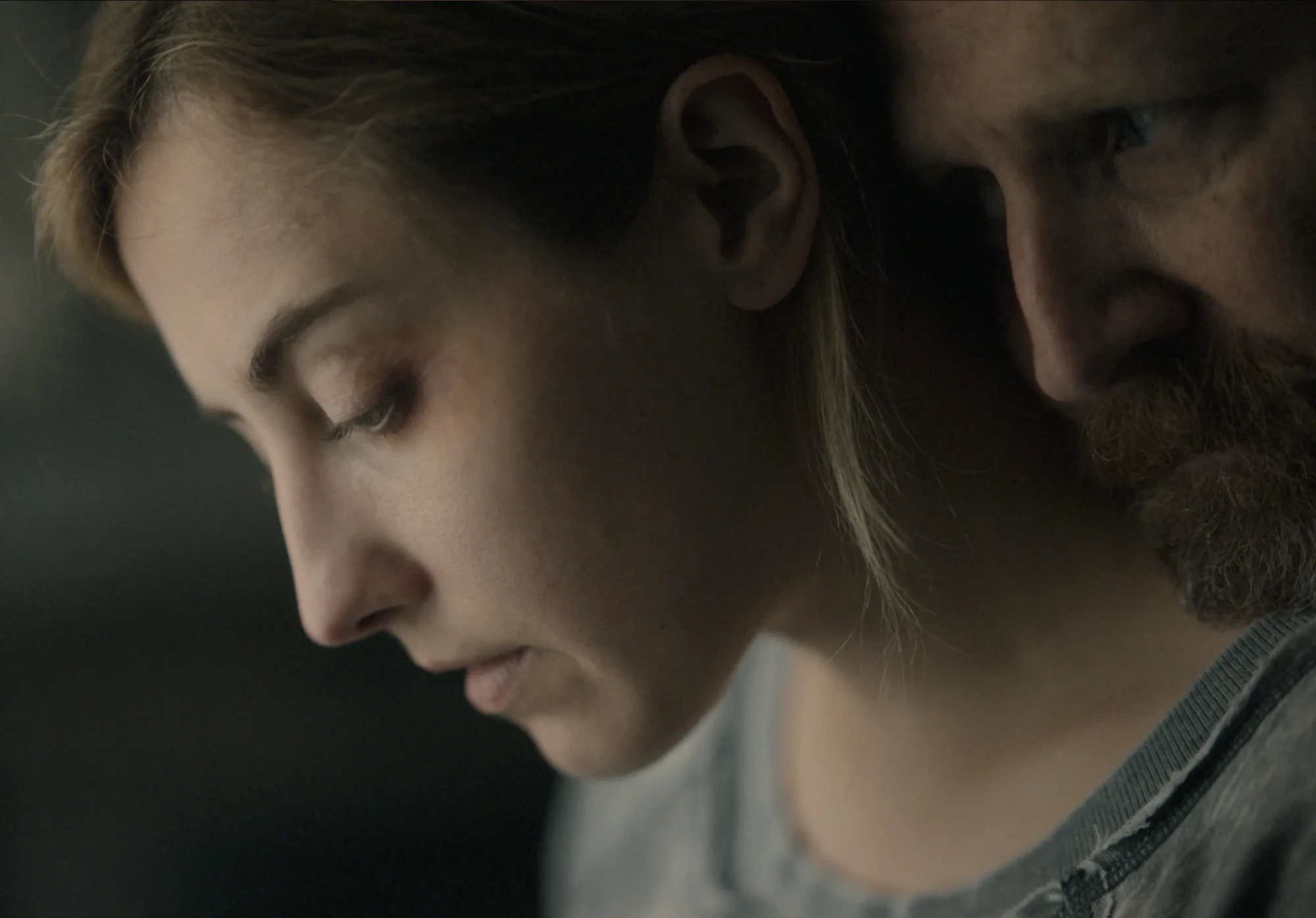 A woman looks down, eyes lowered, as a bearded man leans his head against hers.