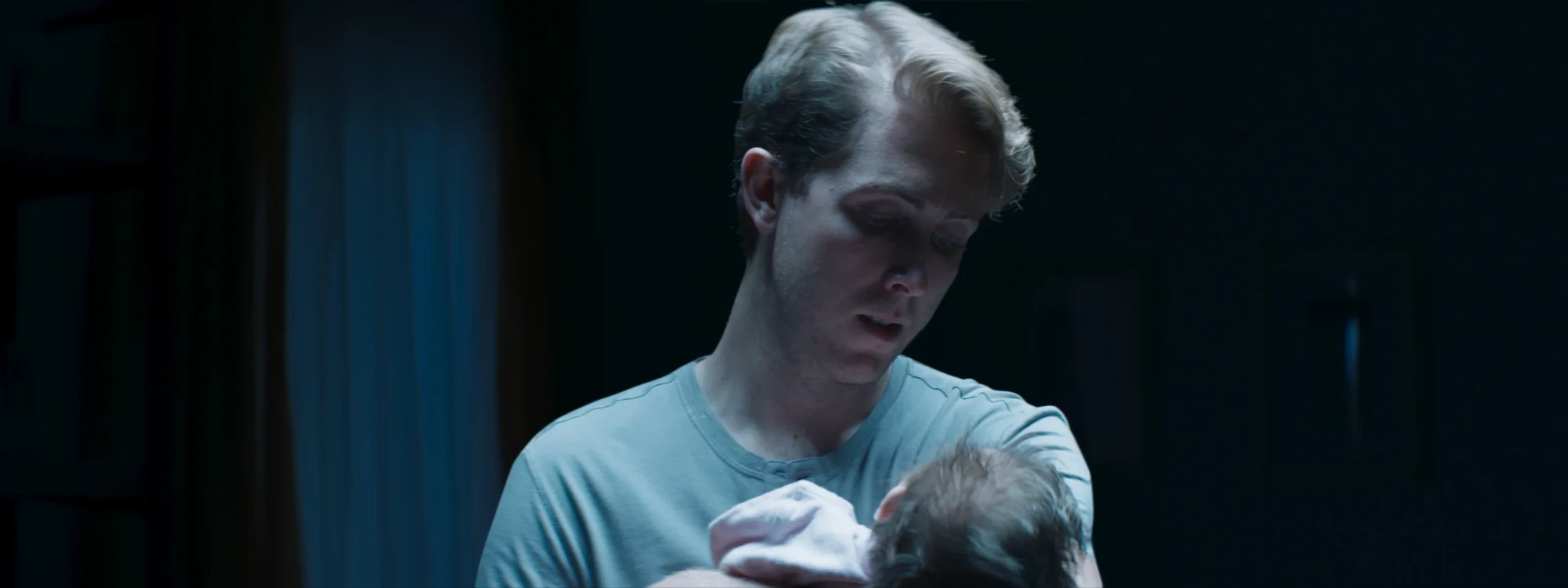 A young father in a T-shirt looks down at the baby in his arms in a dark room.